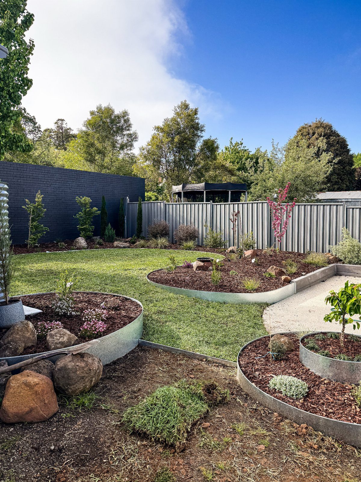 11 Backyard Ideas to Turn Your Outdoor Space Into a Place You Use 11 Landscaped backyard with curved garden beds, metal edging, green grass, and large rocks under a blue sky.