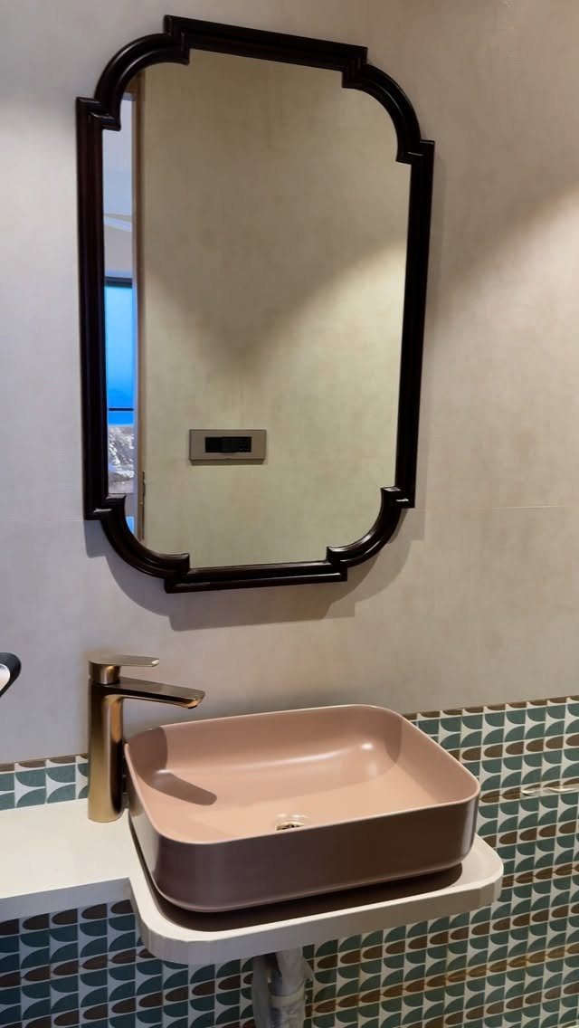 A black framed mirror hangs above a pink vessel sink and gold faucet on a tiled wall.