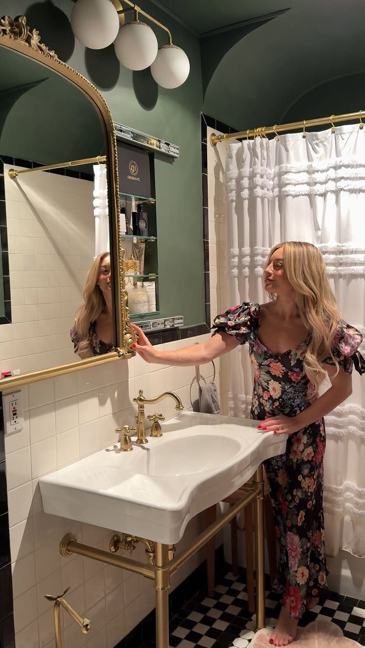 A woman in a floral dress touches a large gold-framed arched mirror in a green bathroom.