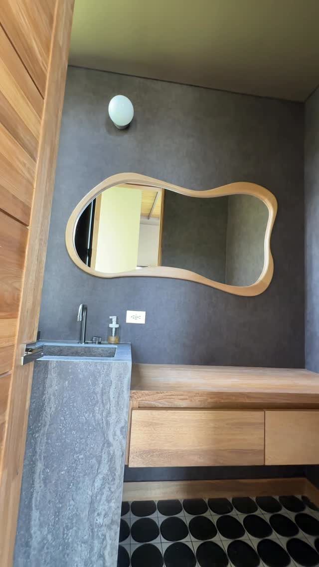 An organic shaped wooden framed mirror mounted on a dark grey bathroom wall.