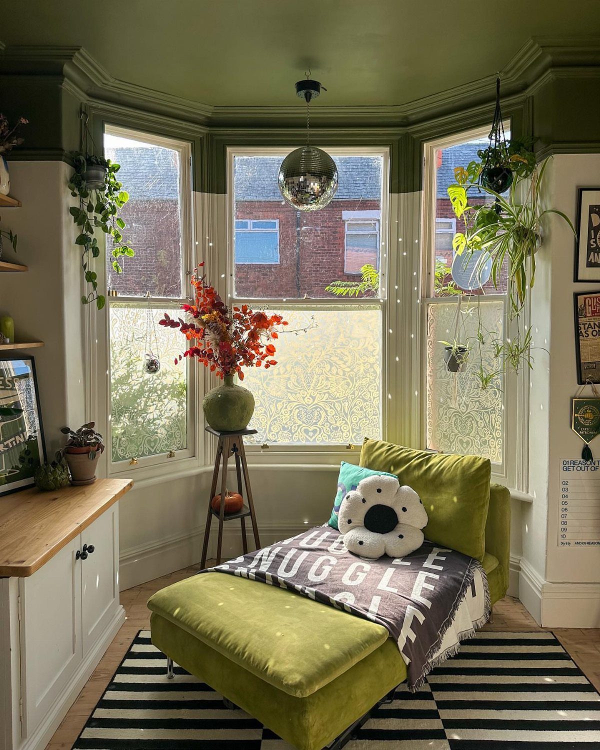 8 Bay Window Ideas for Seating, Dining, Curtains and More 6 A decorated bay window featuring hanging plants, a disco ball, and a green cushioned chaise lounge.