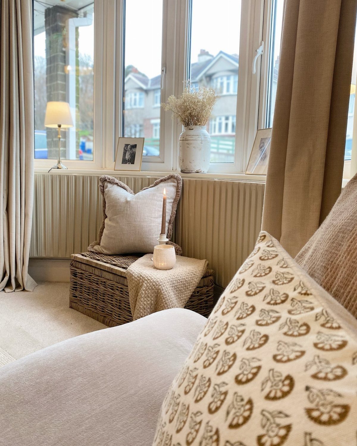 8 Bay Window Ideas for Seating, Dining, Curtains and More 8 Wicker storage ottoman with cushions and a candle placed in a bright bay window setting.