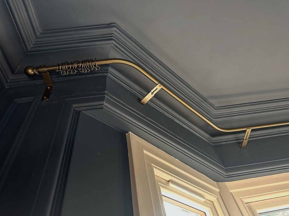 8 Bay Window Treatment Ideas That Balance Light and Privacy 4 A gold curtain rod follows the angled shape of a bay window above dark blue molding.