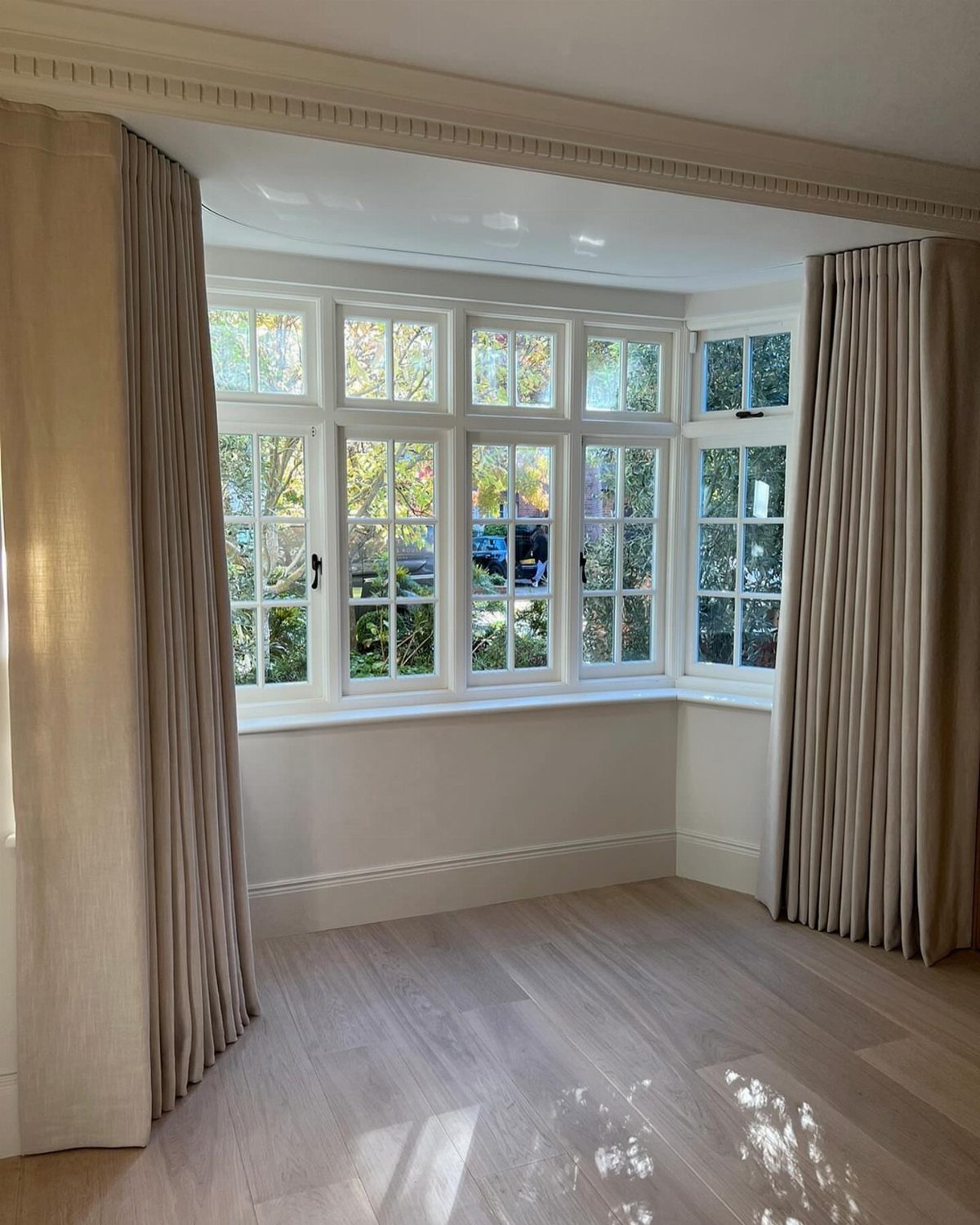 8 Bay Window Treatment Ideas That Balance Light and Privacy 2 Beige floor-length drapery panels framing a large white bay window in a bright room.