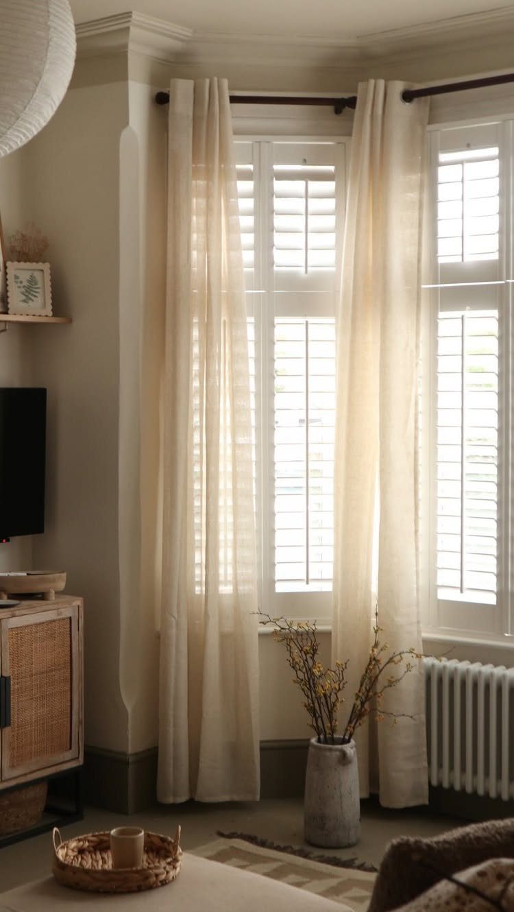 8 Bay Window Treatment Ideas That Balance Light and Privacy 8 Sheer cream curtains hanging in a bay window with white plantation shutters behind them.
