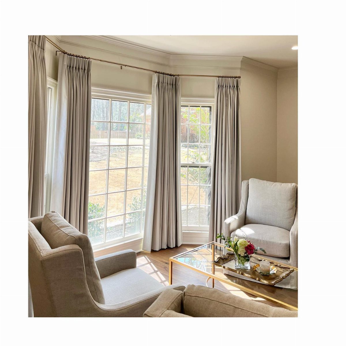 8 Bay Window Treatment Ideas That Balance Light and Privacy 3 Beige drapery panels hanging from a continuous rod across a large bay window in a bright room.