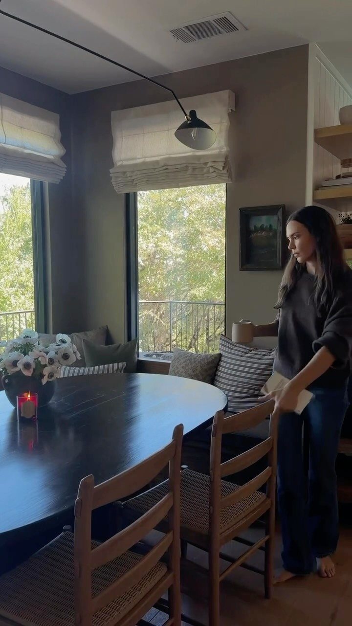 A woman standing near a dining table with various patterned cushions on a window bench.