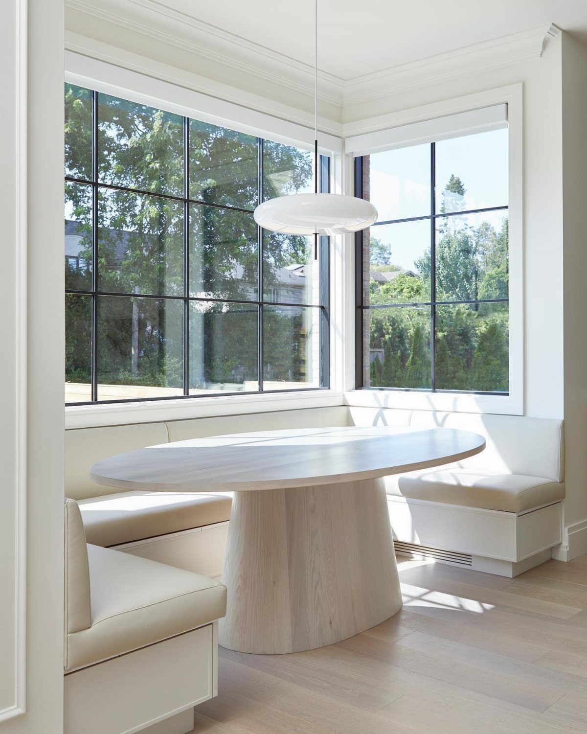An oval wooden pedestal table sits within a white built-in breakfast nook by large windows.