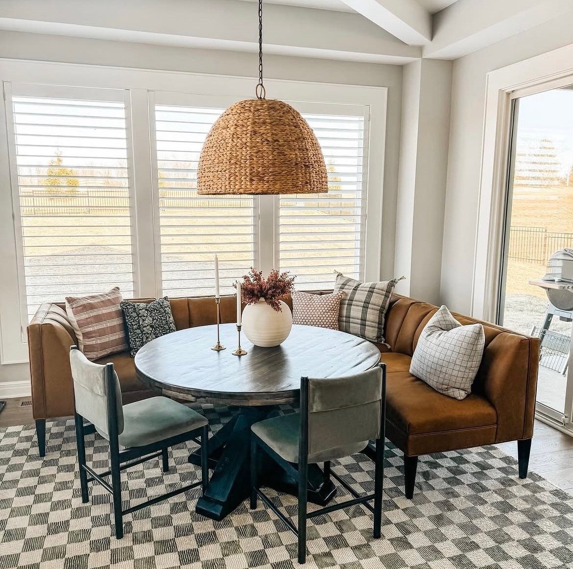 A round wooden table surrounded by a leather banquette and two grey chairs under a woven lamp.