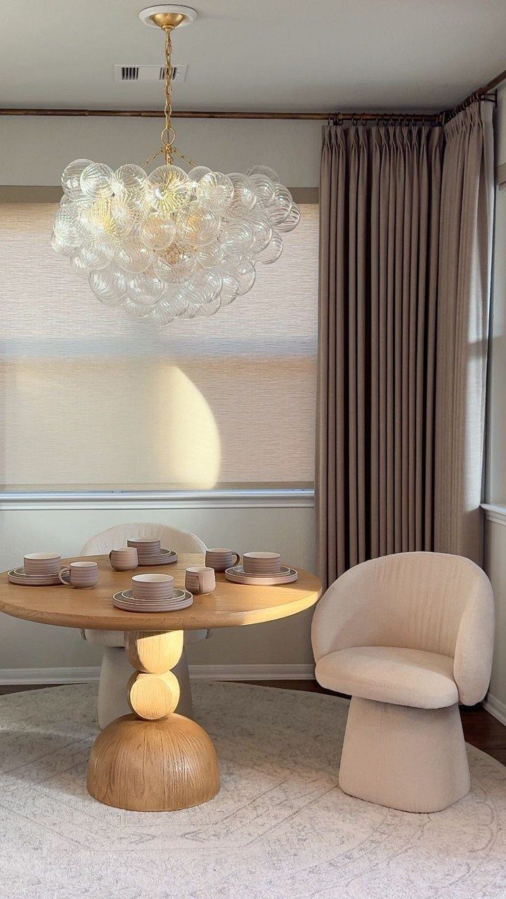 A round wooden pedestal table with coffee cups and a beige armchair under a bubble chandelier.