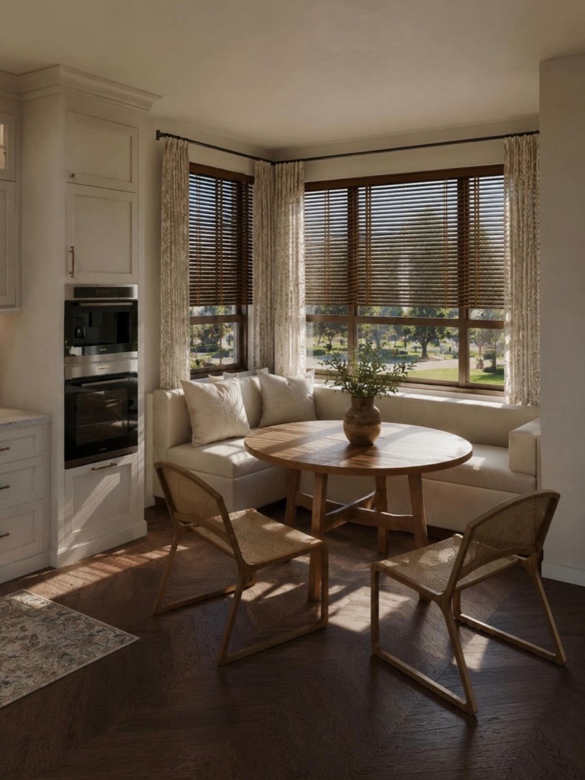 A sunlit breakfast nook with a round wooden table, woven chairs, and a white corner banquette.