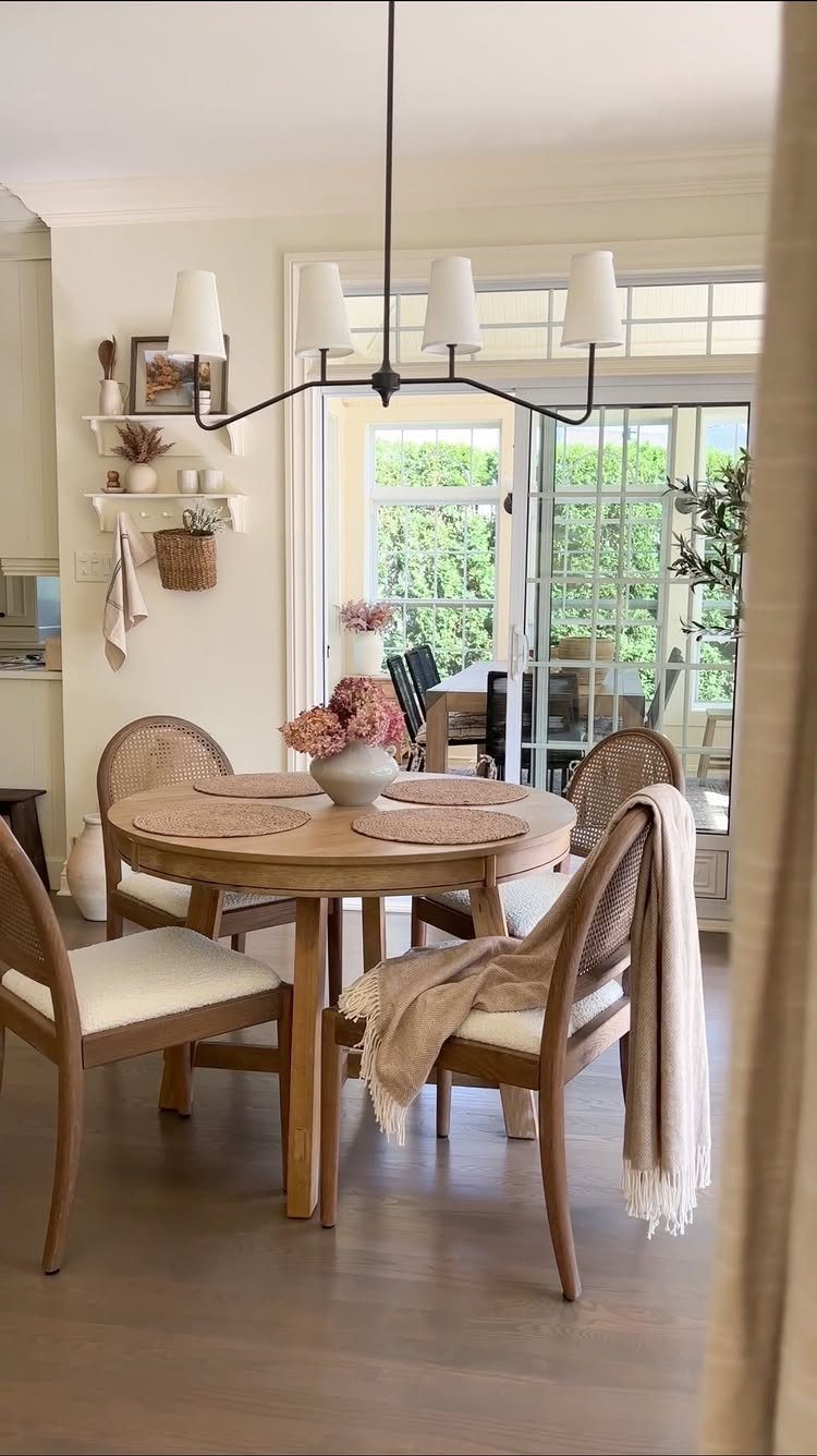 Round wooden dining table with woven chairs and floral centerpiece in a bright breakfast nook.