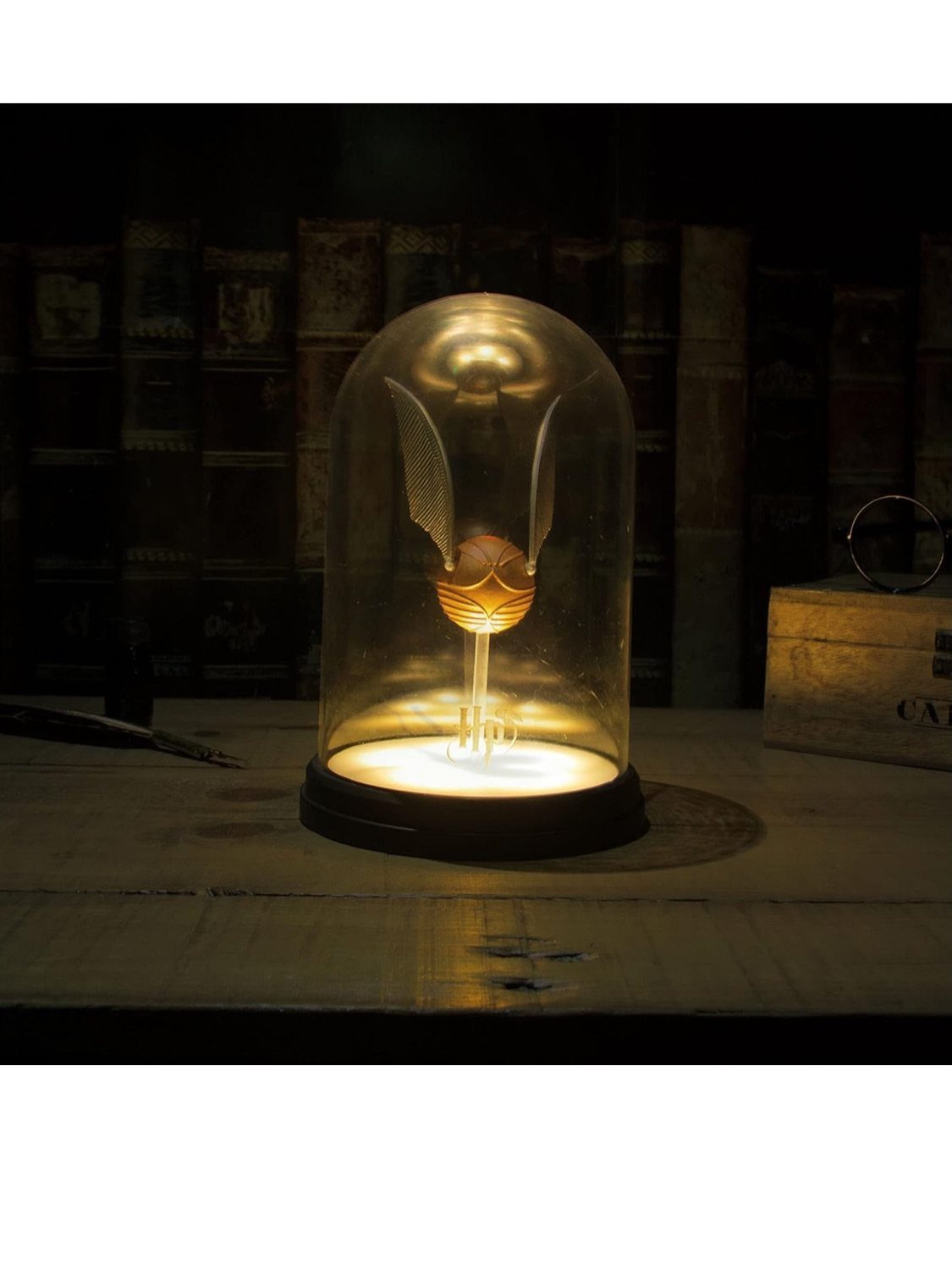 A glowing Golden Snitch lamp with white wings inside a glass cloche on a table.