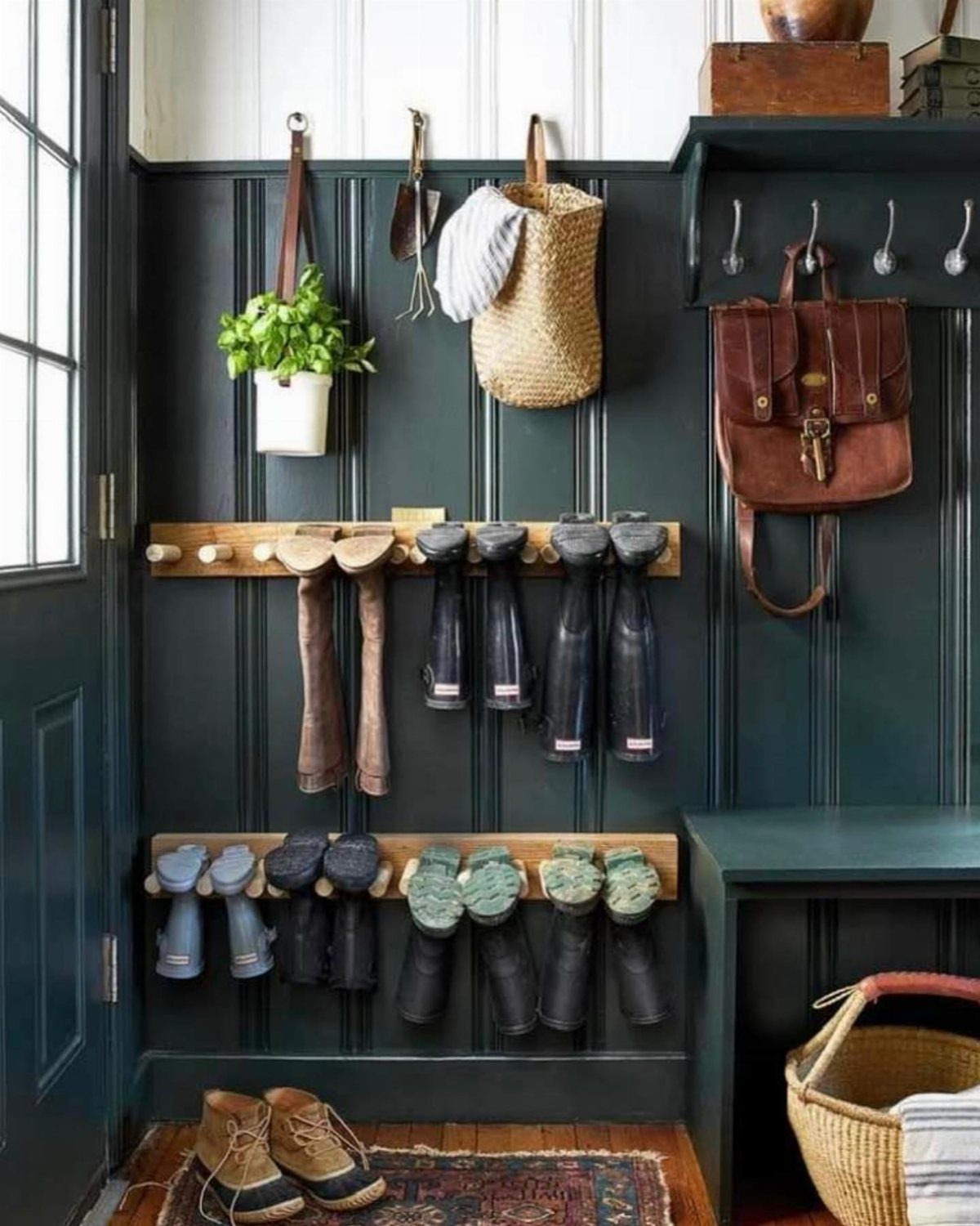 Dark green paneled mudroom wall with wooden boot racks, hanging baskets, and a leather bag.