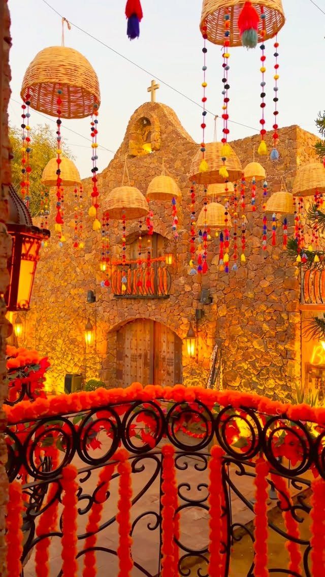 14 Mexican Decor Ideas for Every Room 14 Stone building decorated with colorful hanging baskets and bright orange flower garlands on a wrought iron railing.