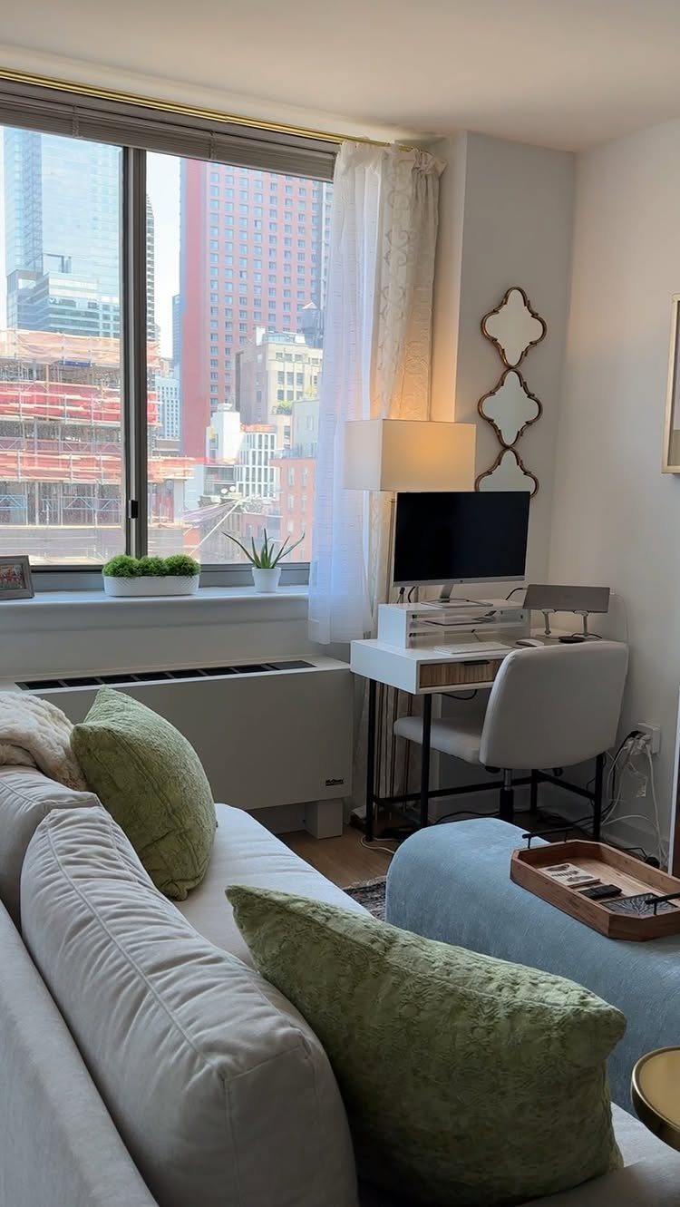Grey sofa with green pillows sits near a white desk and a large city view window.