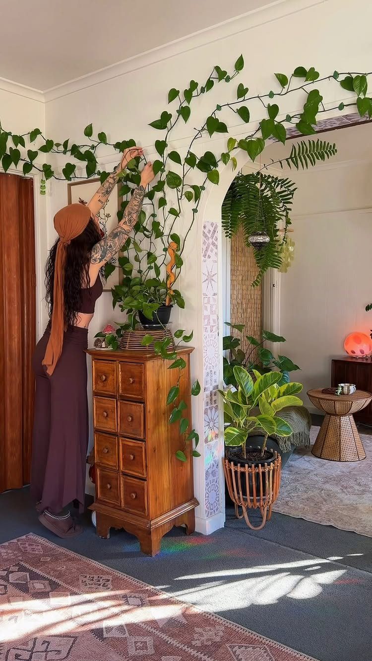A woman tends to climbing green vines growing along a white wall near indoor plants.